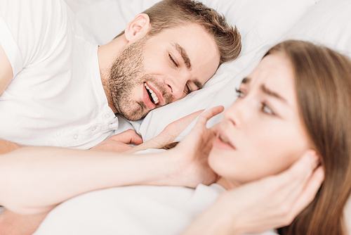 selective focus of exhausted woman  ears with hands while lying in bed near snoring husband