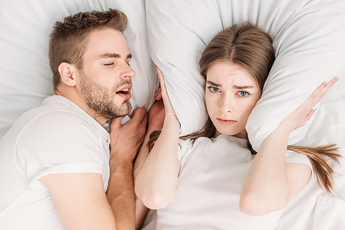 top view of woman  ears with pillow while lying in bed near snoring husband