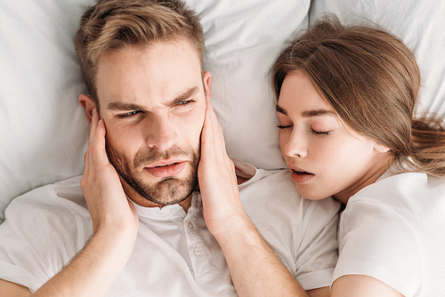 top view of exhausted man  ears with hands while lying in bed near snoring wife