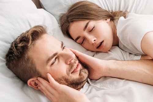 exhausted man  ears with hands while lying in bed near snoring wife