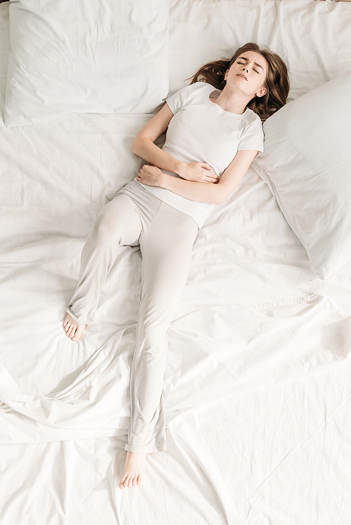 top view of young woman lying in bed with closed eyes and suffering from abdominal pain