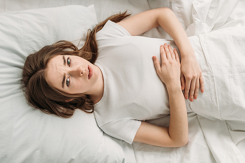 top view of upset girl lying in bed and suffering from stomach pain