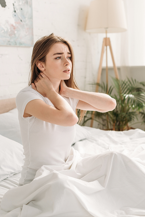 upset girl looking away while sitting in bed and suffering from neck pain