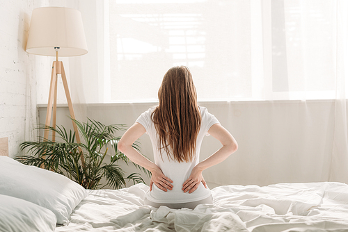 back view of young woman sitting on bed and suffering from back pain