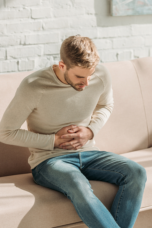 young man sitting on sofa at home and suffering from stomach pain