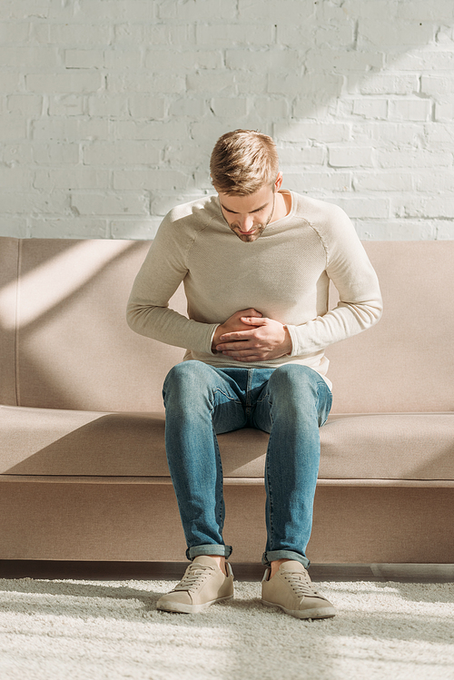 young man sitting on sofa and touching stomach while suffering from abdominal pain