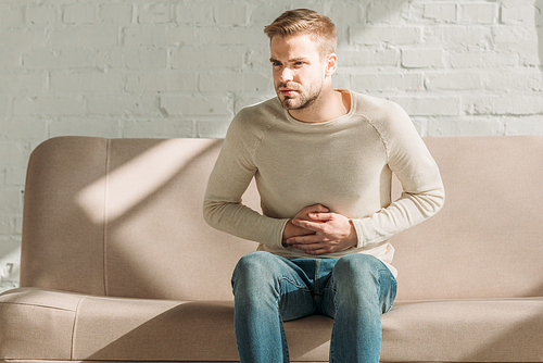 upset man sitting on sofa and touching stomach while suffering from abdominal pain