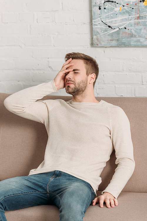 young man sitting on sofa with closed eyes while suffering from migraine