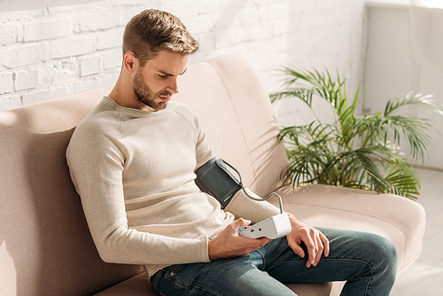 handsome man sitting on sofa at home and measuring blood pressure with tonometer