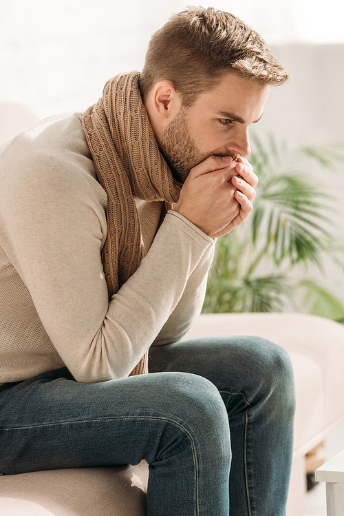 sick man in warm scarf coughing in folded hands while sitting on sofa