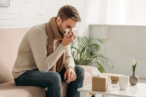 ill man sneezing in napkin while sitting on sofa near table