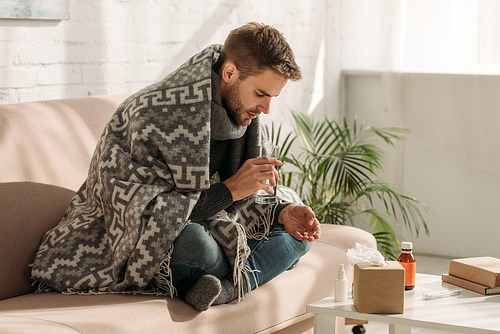 sick, unhappy man, wrapped in blanket, sitting on sofa and taking medicines