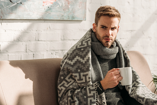 sick, unhappy man, wrapped in blanket, looking away while holding cup of warming drink