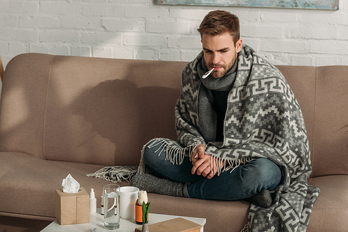 upset, diseased man measuring temperature while sitting on sofa near table with medicines