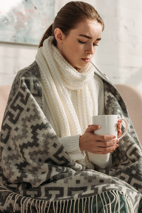 sick woman with warm scarf around neck, wrapped in blanket, holding cup of tea