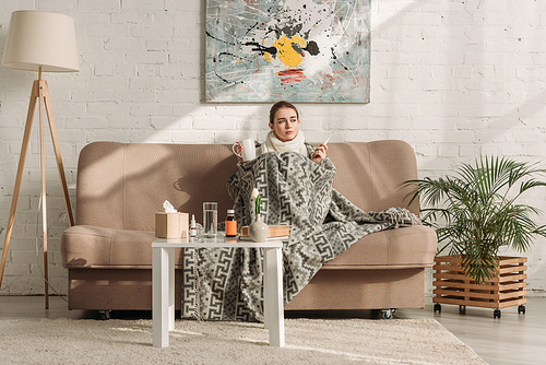 diseased woman wrapping in blanket while sitting on sofa and holding thermometer and cup of warming drink