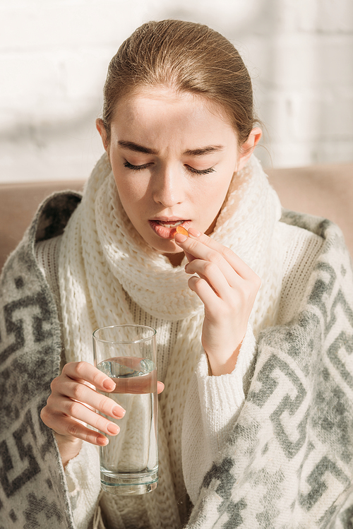 sick girl, wrapped in blanket, holding glass of water while taking medicine