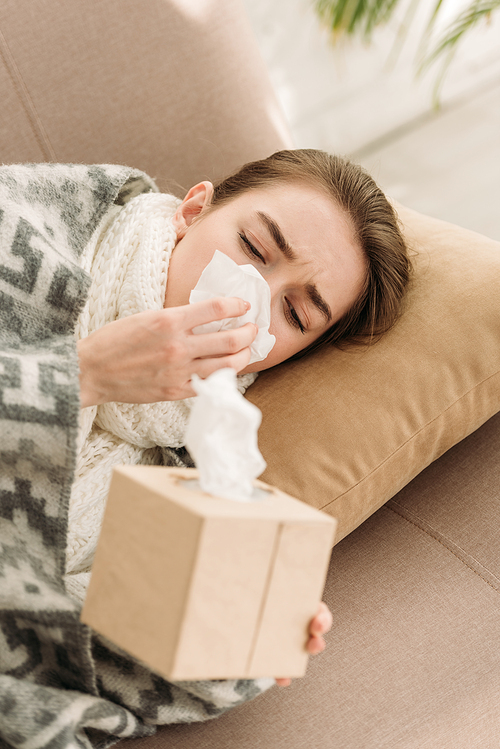 sick girl, covered with blanket, lying on sofa and sneezing in napkin