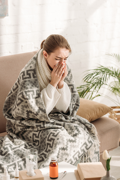 diseased woman, wrapped in blanket, sitting on sofa and sneezing in napkin