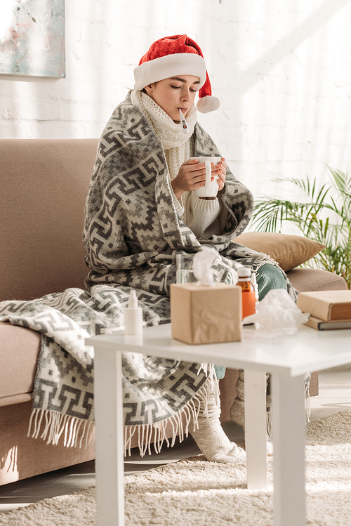 sick woman in santa hat, wrapped in blanket, measuring temperature and holding cup of warming drink