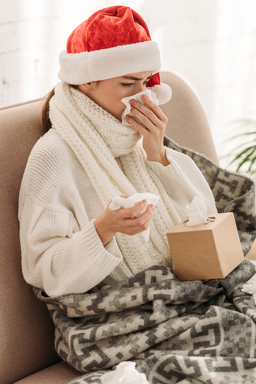 sick woman sneezing in napking while sitting on sofa in santa hat