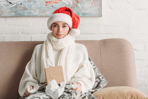 sick woman in santa hat and warm scarf sitting on sofa, holding napkins and