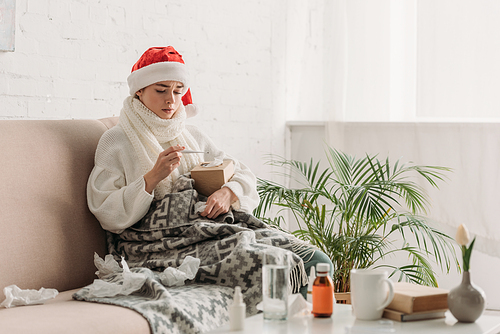 sick woman in santa hat, wrapped in blanket, sitting on sofa near table with medicines and looking at thermometer