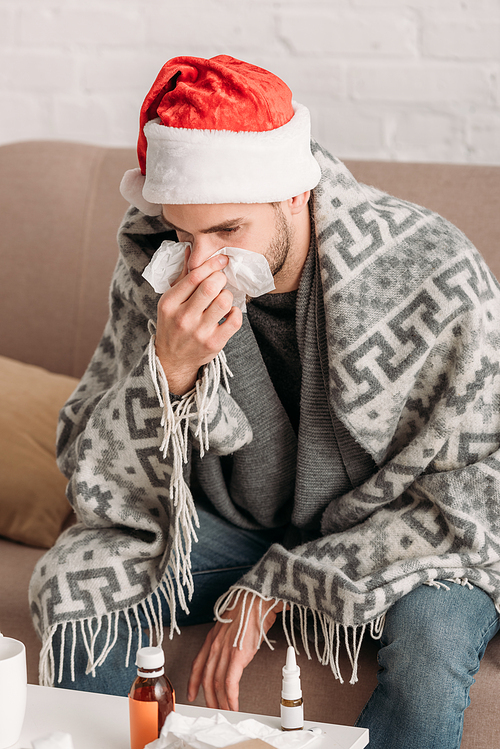 sick man sitting in santa hat, wrapped in blanket, sitting in sofa and sneezing in napkin