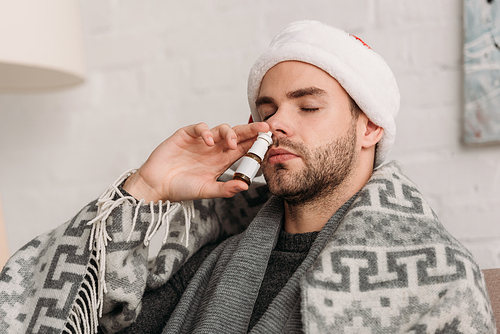 unhealthy man in santa hat, wrapped in blanket, using nasal spray with closed eyes