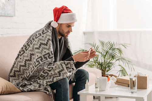 sick man in santa hat, wrapped in blanket, sitting near table with medicines and holding thermometer