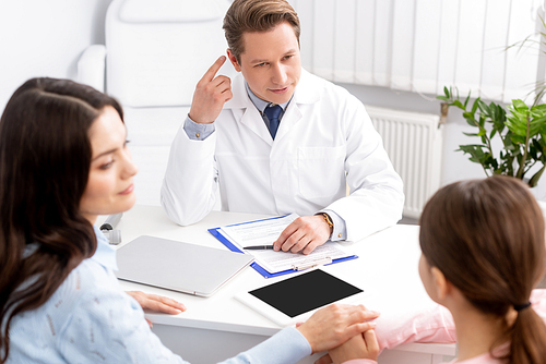 ent physician pointing with finger at ear near mother and daughter sitting at desk
