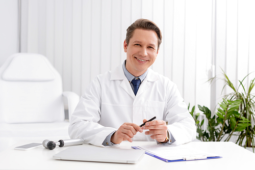 smiling otolaryngologist  while sitting ant workplace near otoscope, laptop and clipboard