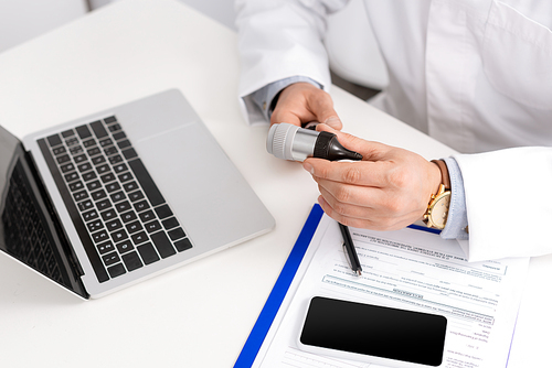 cropped view of otolaryngologist holding otoscope near clipboard, laptop and smartphone with blank screen