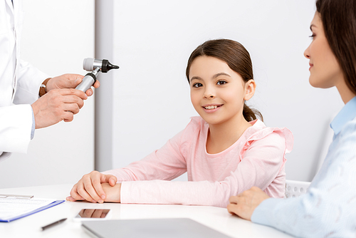 cropped view of otolaryngologist holding otoscope near smiling child and mother