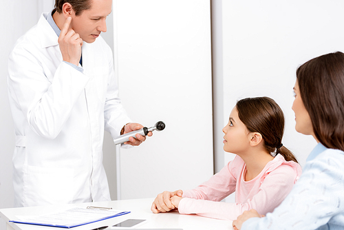 mother and daughter looking at otolaryngologist holding otoscope and pointing with finger at his ear