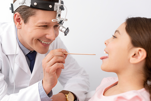 cheerful ent physician examining throat of cute child with tongue depressor