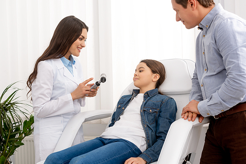 smiling ent physician pointing with finger at otoscope near smiling kid sitting in medical chair and her father standing nearby