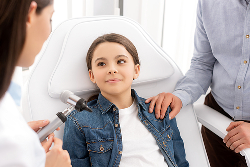 cropped view of father touching shoulder of daughter sitting in medical chair near otolaryngologist with otoscope