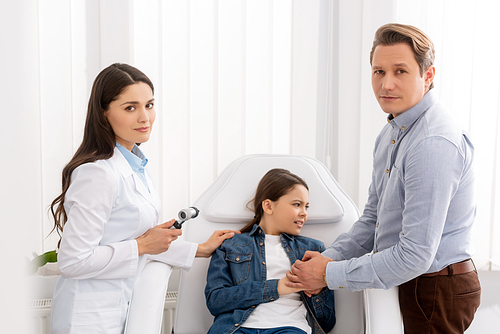 father holding hands of worried daughter sitting in medical chair near ent physician with otoscope