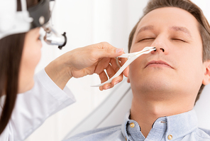 cropped view of otolaryngologist examining nose of handsome man with nasal speculum