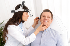 attractive smiling otolaryngologist in ent headlight examining ear of cheerful man