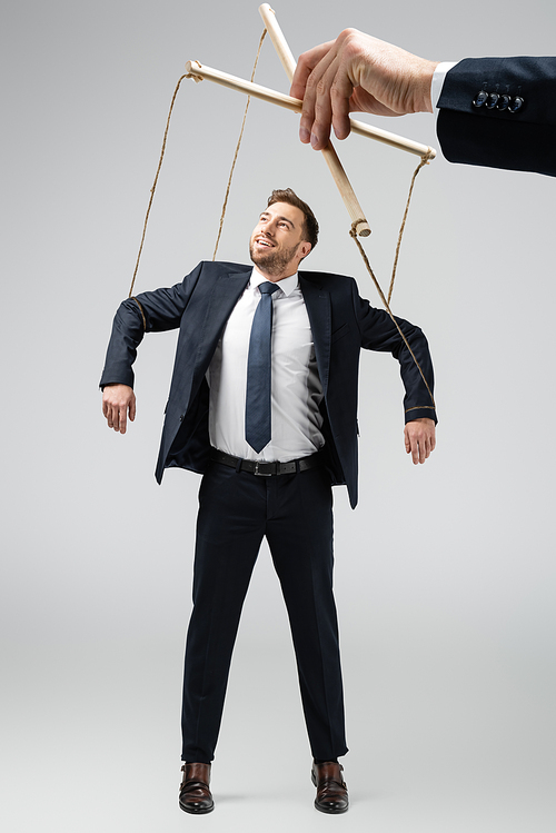 cropped view of puppeteer holding smiling businessman marionette on strings isolated on grey