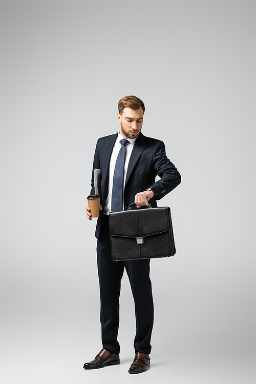 businessman with leather suitcase and paper cup looking at wristwatch isolated on grey