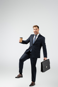 cheerful businessman with leather suitcase and paper cup isolated on grey