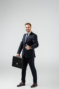 smiling businessman with leather suitcase showing thumb up isolated on grey