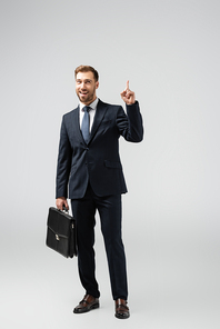 smiling businessman with leather suitcase pointing up isolated on grey