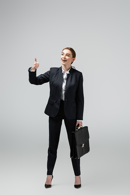 cheerful young businesswoman with leather suitcase showing thumb up isolated on grey