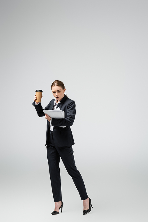shocked young businesswoman with coffee to go and papers isolated on grey