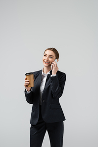 happy young businesswoman with coffee to go talking on smartphone isolated on grey