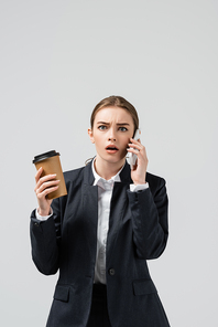 shocked young businesswoman with coffee to go talking on smartphone isolated on grey